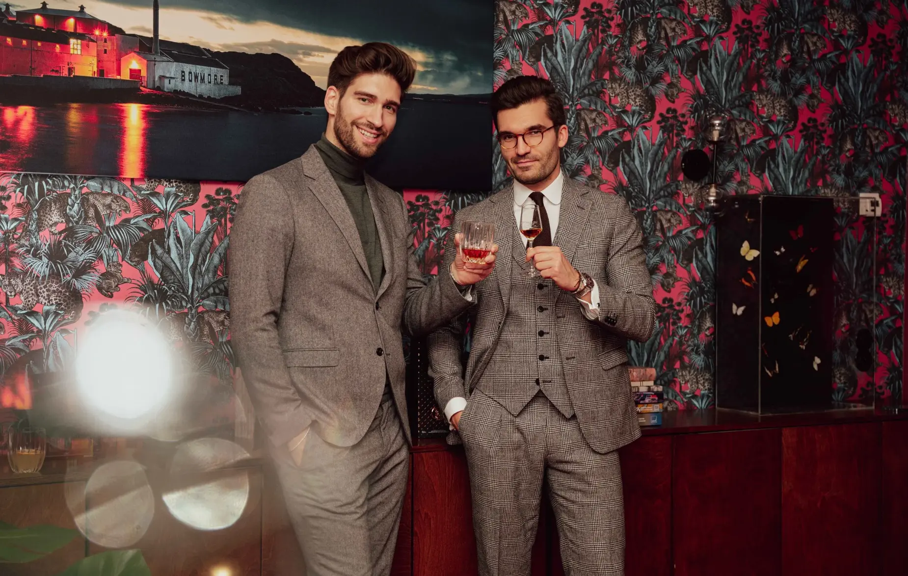 Two guests in tailored suits raising whisky glasses in front of a BOWMORE distillery artwork at the members' club
