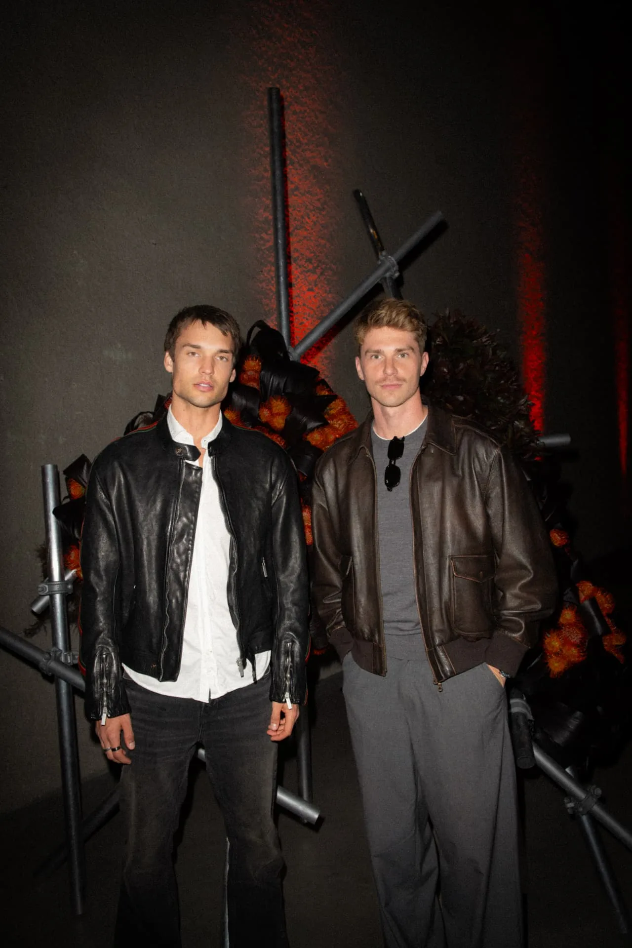 Two guests in leather jackets before the red-lit floral backdrop at the EX NIHILO event