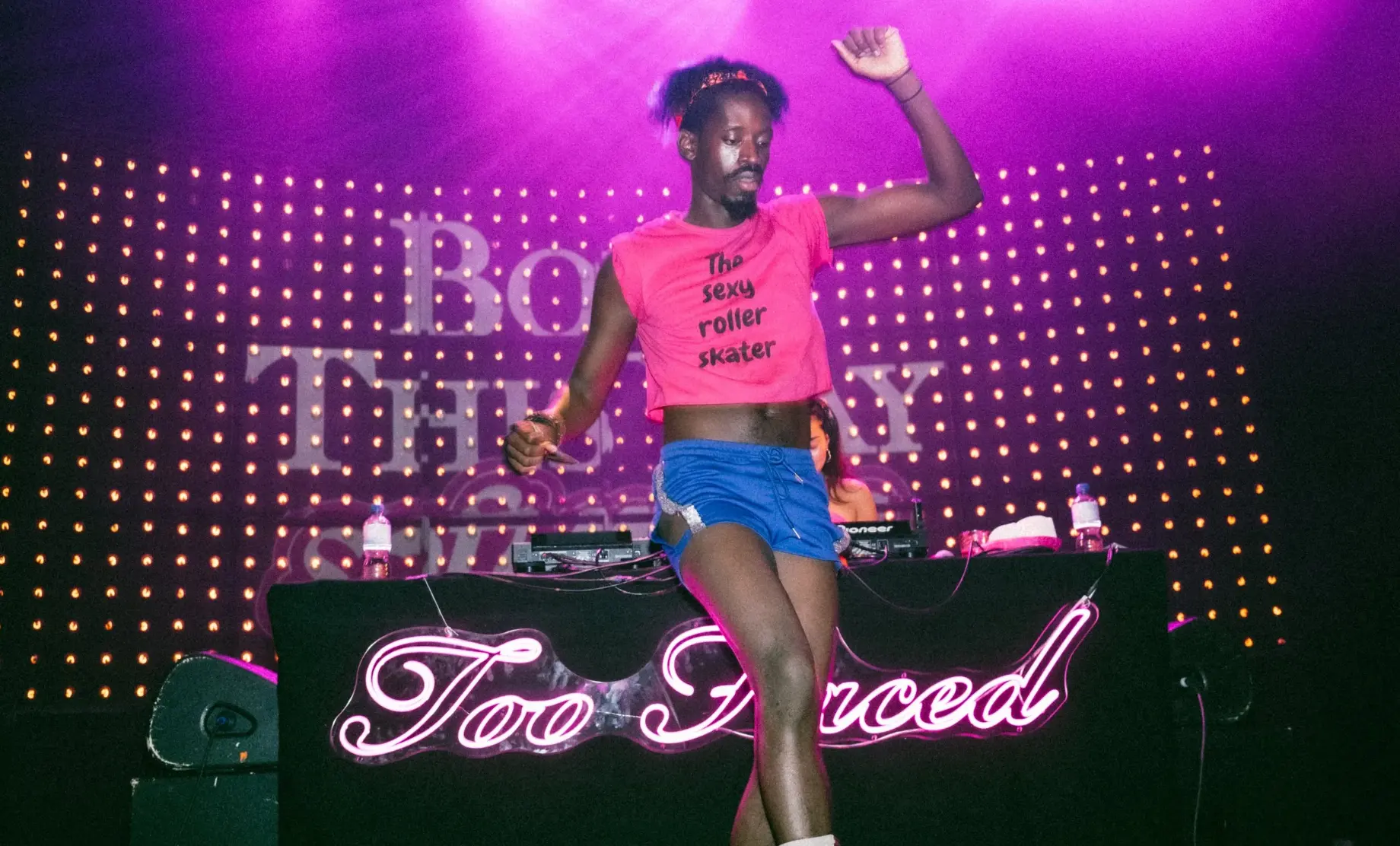 Roller skater dancing on the DJ booth in front of the glowing pink too faced neon sign